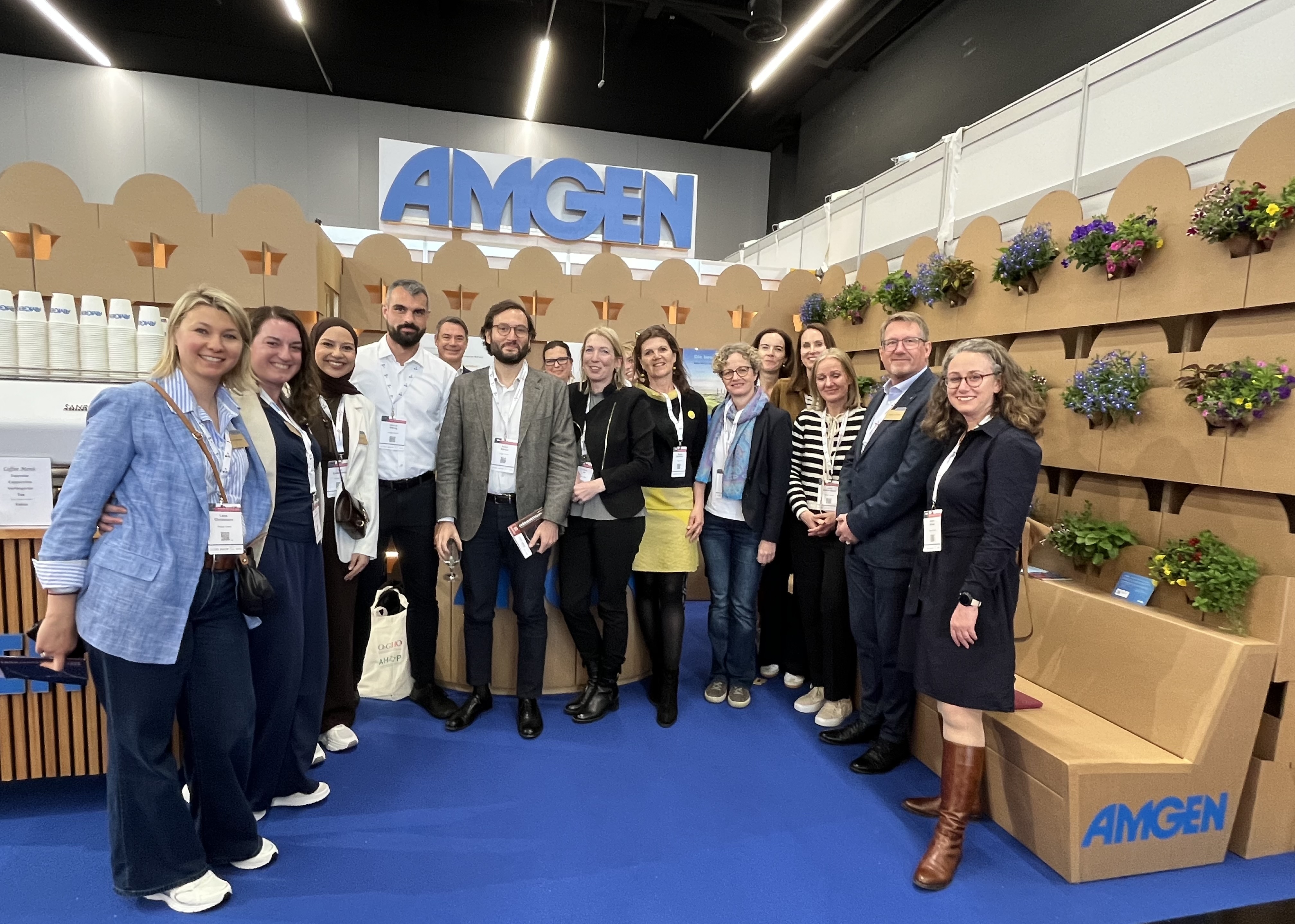 Gruppe von Menschen vor einem AMGEN-Messestand mit Kartonelementen und Blumenwand, alle tragen Namensschilder und lächeln in die Kamera.