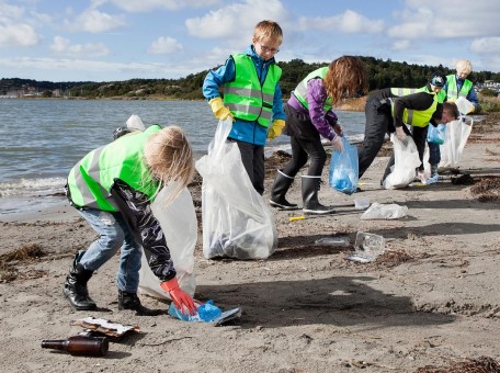 Eine Gruppe von Erwachsenen und Kindern in grünen Warnwesten sammeln an einem Strand Müll auf
